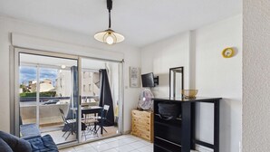 A modern living room with a sofa, a dining table with chairs, a TV, a fan, and a sliding glass door leading to a balcony. - Cozy Studio Apartment with Pool Access near Beach in Marseillan, Sleeps 4 (Marseillan-Plage)
