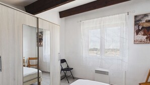A bedroom with a bed, a chair, a wardrobe, a window with curtains, and a radiator. - Charming Studio with Mezzanine & Terrace for 6 in Marseillan, Parking Included (Marseillan-Plage)
