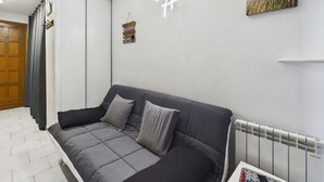 A modern living room with a grey sofa, a radiator, a wall-mounted artwork, and a door with a curtain. - Charming Studio with Mezzanine & Terrace for 6 in Marseillan, Parking Included (Marseillan-Plage)