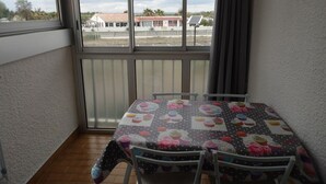 Un balcon avec une table recouverte d’une nappe à motifs de cupcakes, deux chaises et une vue sur des immeubles, avec un panneau solaire en arrière-plan.