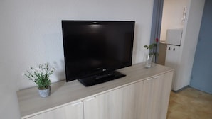 Un téléviseur à écran plat est posé sur un meuble en bois de couleur claire, dans une pièce dont le mur est blanc et où l’on aperçoit une porte au fond.
