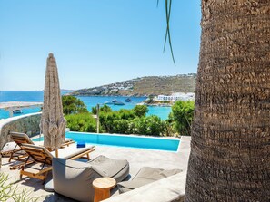 Un espace piscine avec des chaises longues et un parasol, offrant une vue sur une ville côtière avec ses bateaux et ses bâtiments.