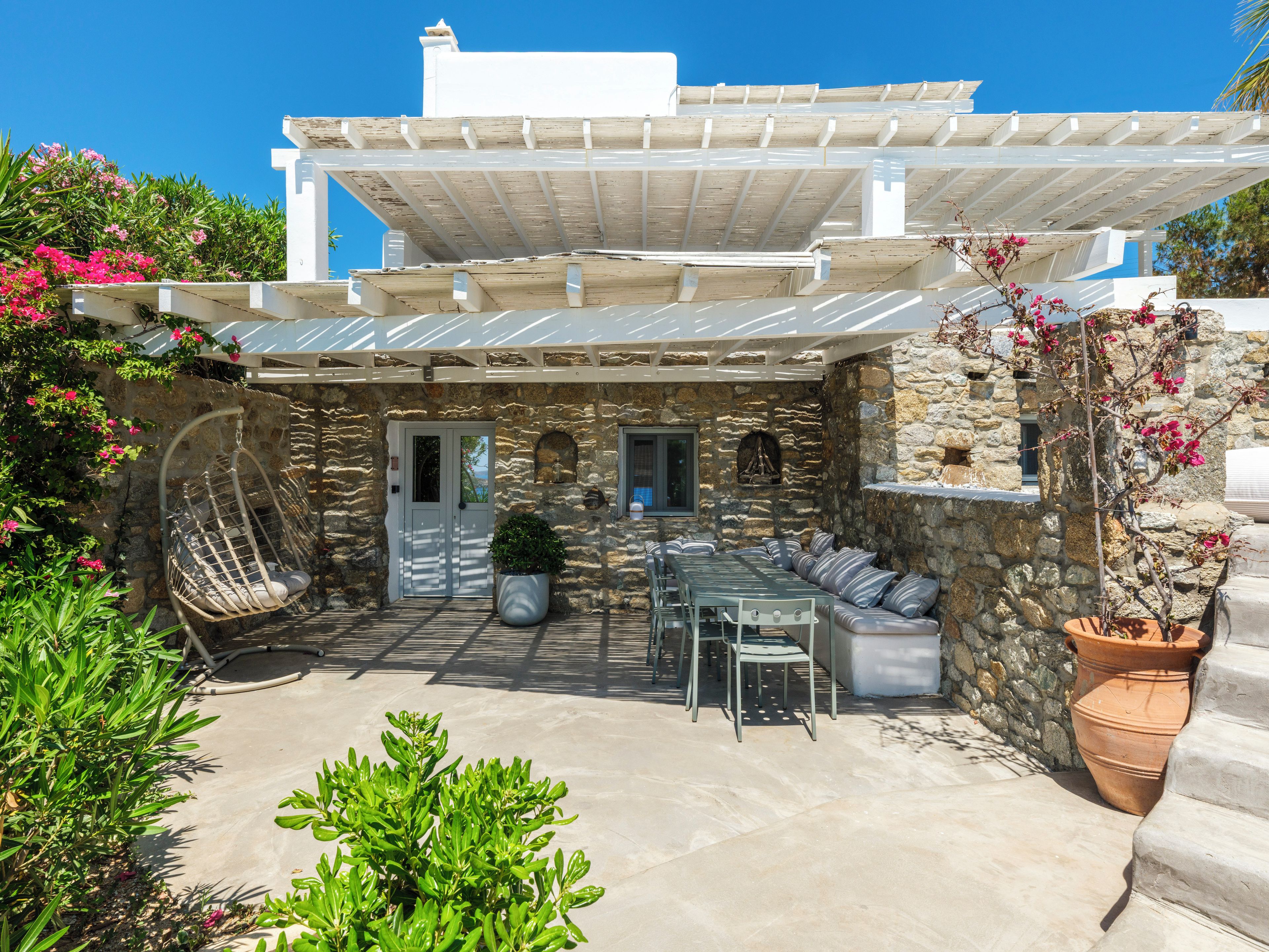 A stone building with a white door, a hanging chair, a table with chairs, and a pergola.