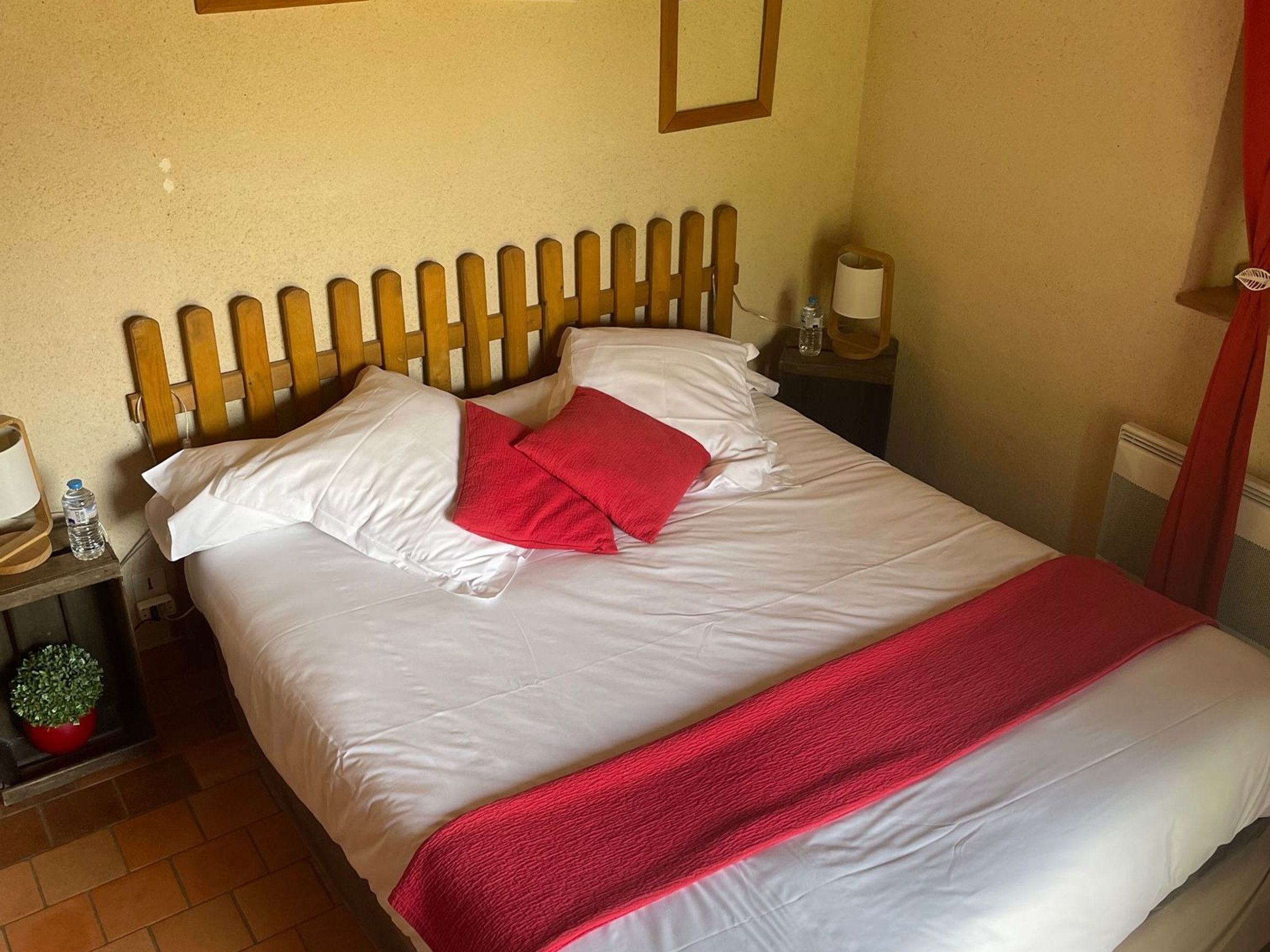 Una cama bien hecha con sábanas blancas y una manta roja, un cabecero de madera, una mesita de noche con una lámpara y una pequeña planta en maceta.
