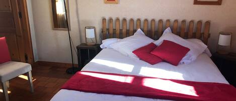 Una cama bien hecha con ropa de cama blanca y roja, una lámpara de noche y una silla con un cojín rojo.