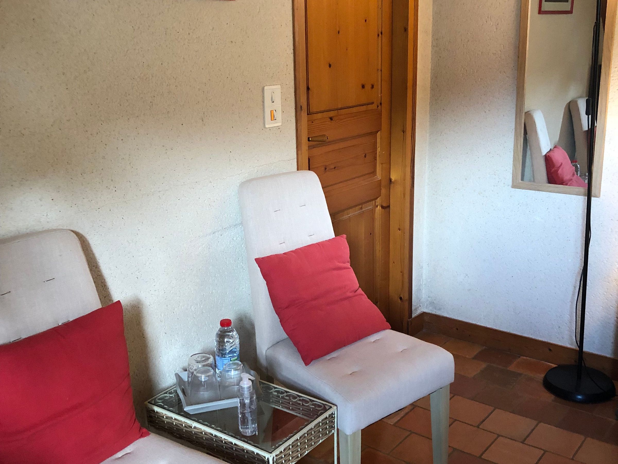 Een kamer met twee witte stoelen, elk voorzien van een rode kussen, een klein tafeltje met een waterfles en glazen, en een vloerlamp.