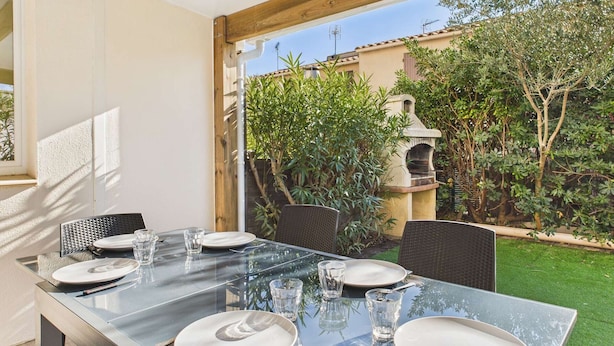 A dining area with a glass table set for four, surrounded by wicker chairs, and a view of a garden with a stone fountain and greenery.