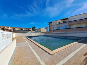 En swimmingpool under en klar blå himmel, omgivet af bygninger med altaner og et hvidt hegn.