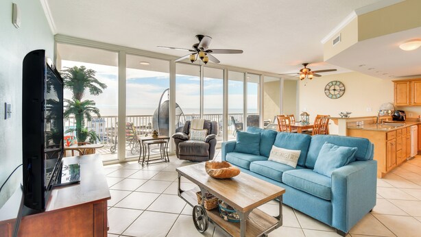 A spacious living area with a blue sofa, a wooden coffee table, and a television on a stand. There is a balcony with a view of palm trees and a cityscape. - Gulf Shores Beachfront Condo w/ Community Pool! (Gulf Shores)