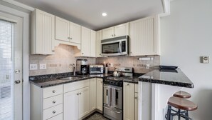 A compact kitchen with white cabinets, a built-in microwave, a stove, and a coffee maker. There are two stools at the counter. - Walk to Commercial Street! Couples Retreat w/ Deck (Provincetown)