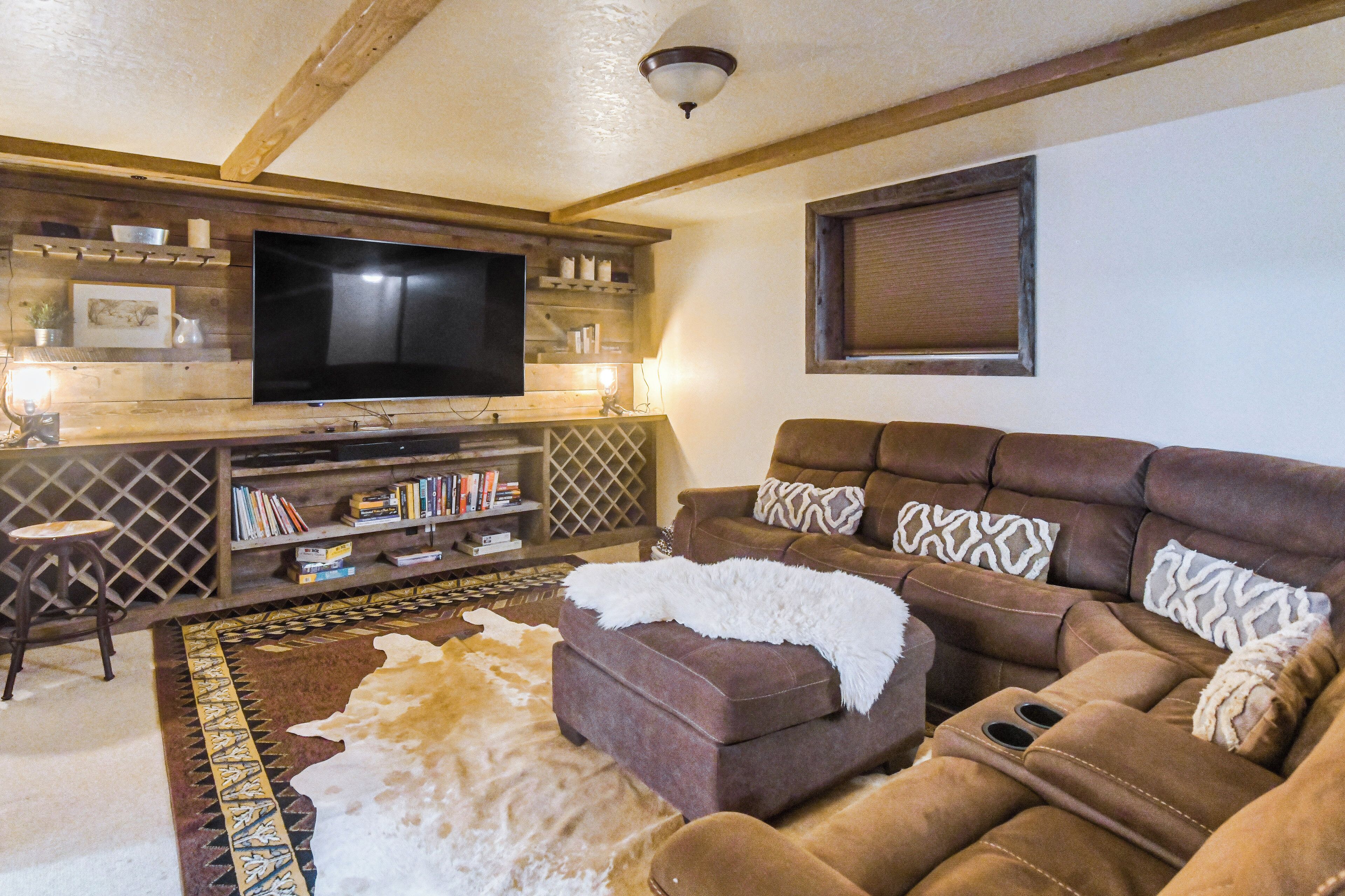 A cozy living room with a large flat-screen TV, a brown leather sofa, a patterned rug, and a wooden entertainment unit with books and decorative items.