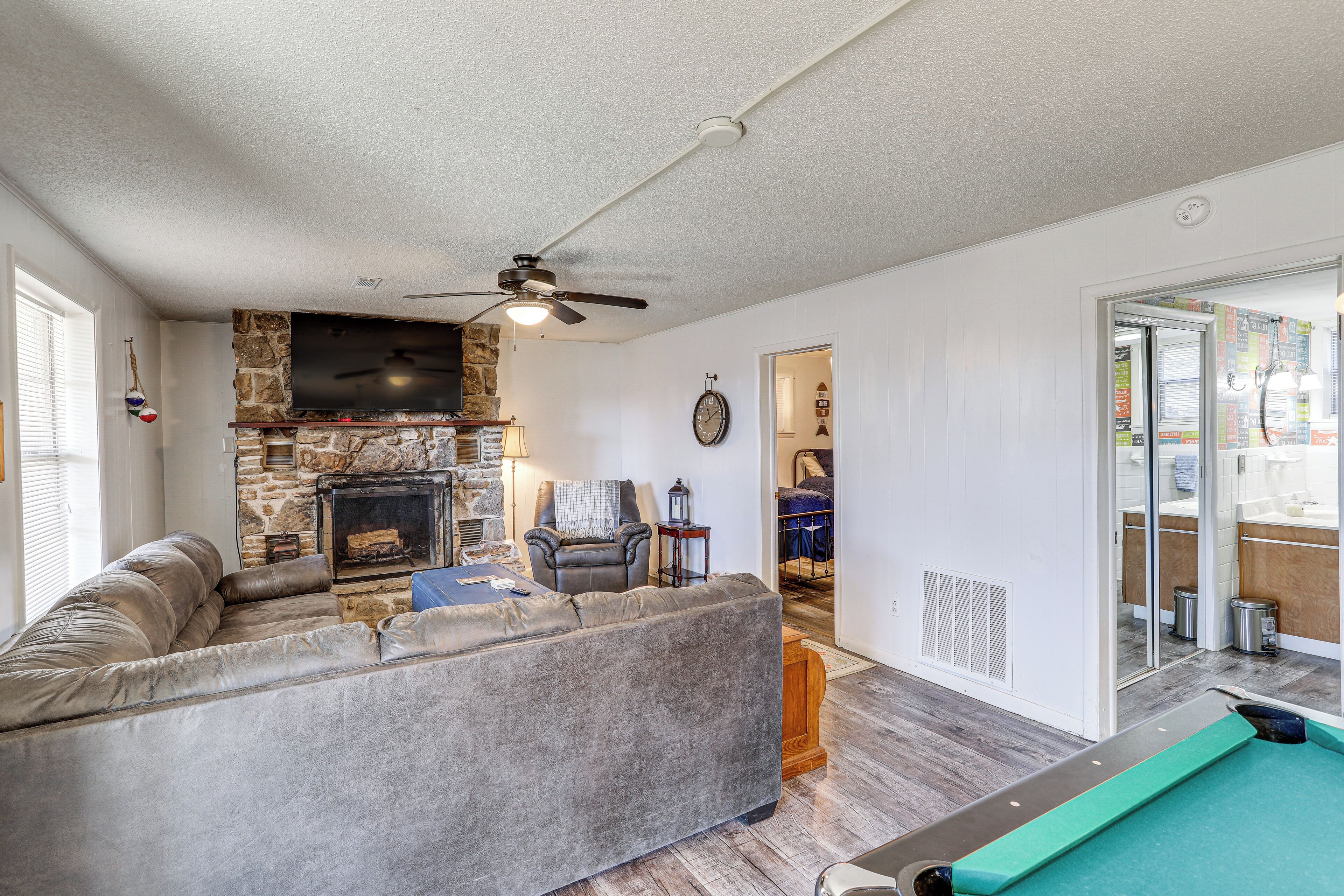 A living room with a stone fireplace, a grey sofa, a ceiling fan, and a pool table.