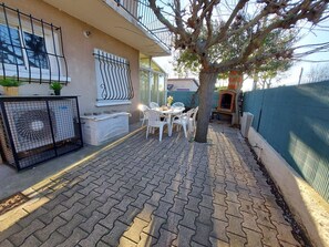 Une terrasse avec un sol en briques, une table blanche avec des chaises, un petit arbre et un espace délimité par une clôture, où se trouve un barbecue.