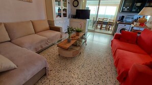 A living room with a beige sofa, a red sofa, a coffee table, and a TV. There is a kitchen area with a washing machine and a dining area with a table and chairs.