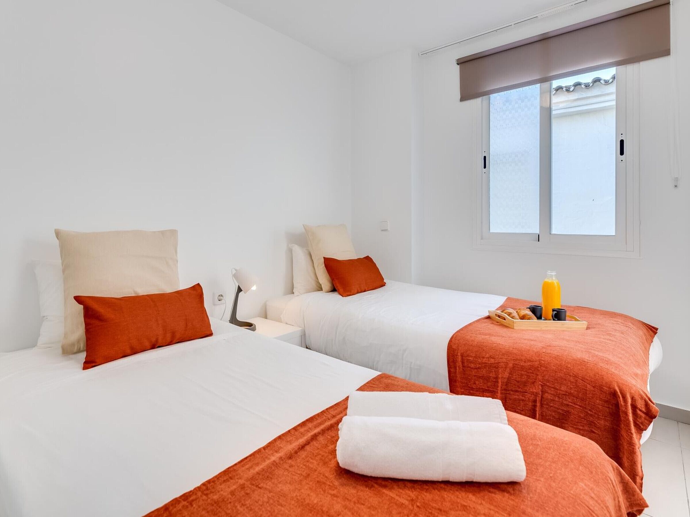 A hotel room with two beds, each with white linens and orange accents, a bedside table with a lamp, and a window with a view.