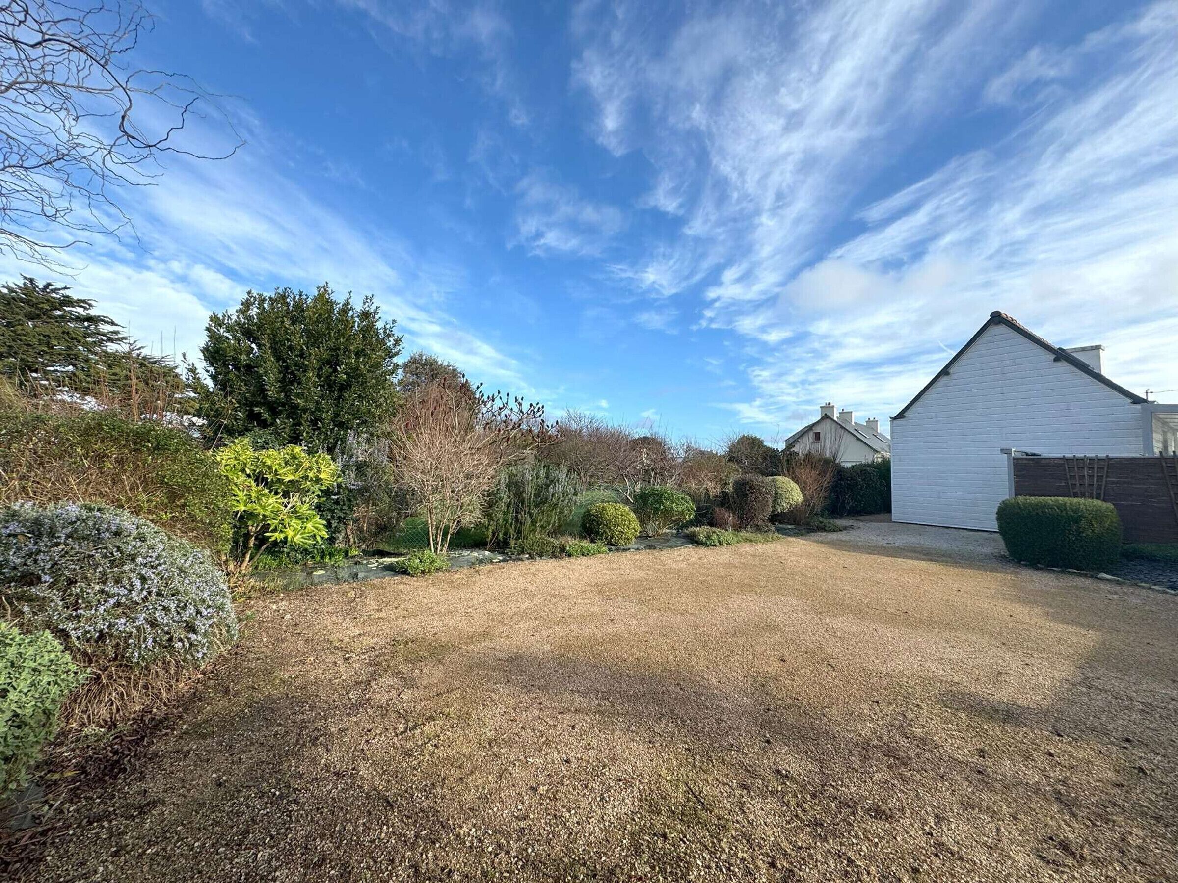 A residential property with a gravel driveway, a white garage, and a variety of shrubs and trees.