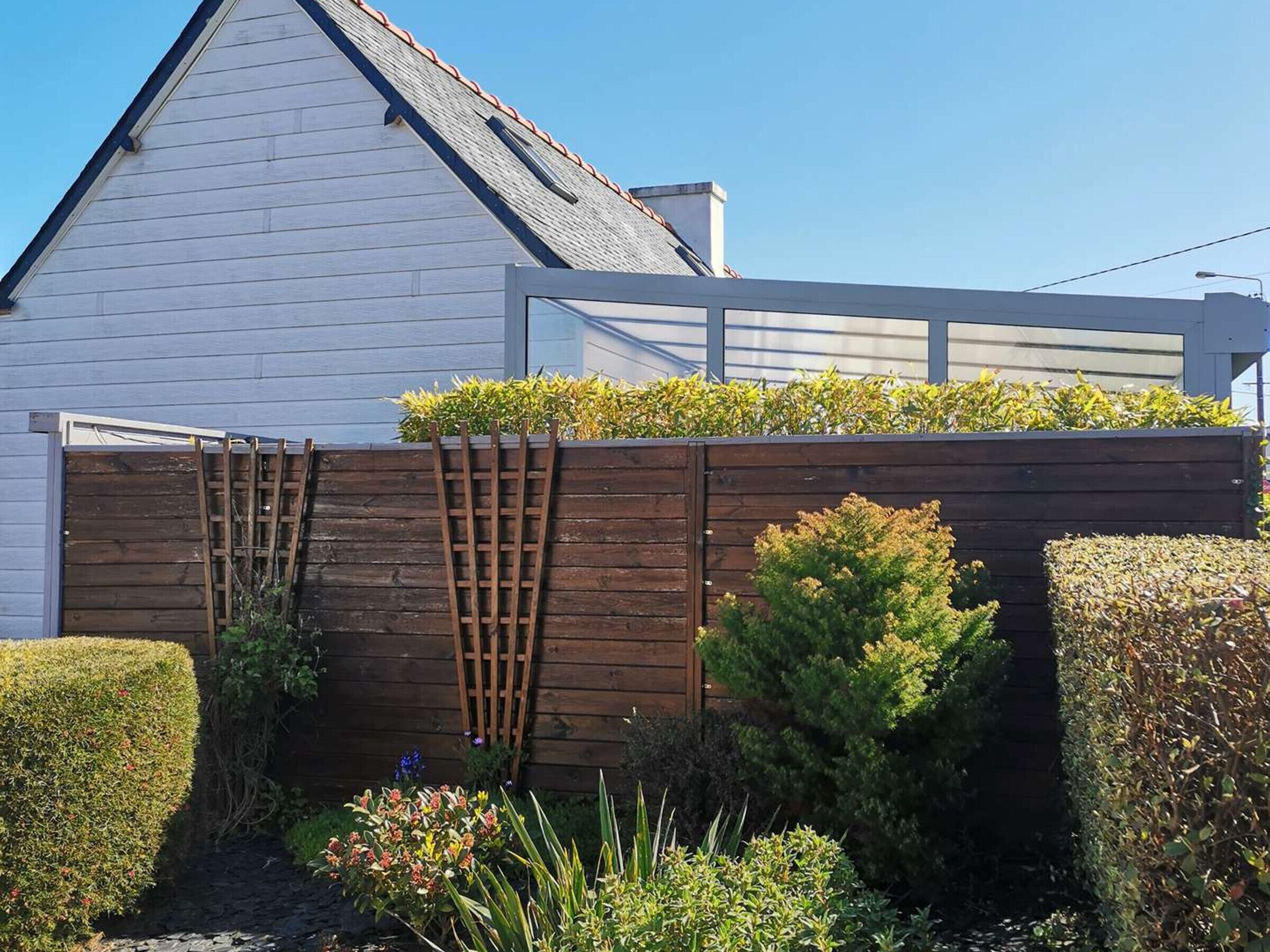 A white house with a sloped roof, a wooden fence, and various shrubs and plants.