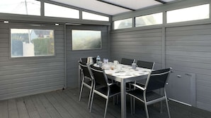 A covered patio area with a table set for four, surrounded by metal chairs, large windows, and grey walls. - Seaside House with WIFI, Veranda, and Parking – 320m from the Beach in Trégastel (Trégastel)