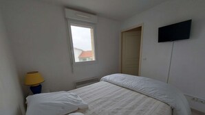 A bedroom with a bed, a window, a radiator, a lamp, and a TV mounted on the wall. - Charming 2-Room Cabin Apartment, 50m from Beach, A/C, Parking (Marseillan-Plage)