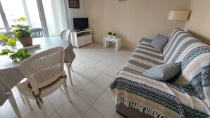 A living room with a sofa, a TV, a dining table with chairs, and a small table with a lamp. - Charming 2-Room Cabin Apartment, 50m from Beach, A/C, Parking (Marseillan-Plage)