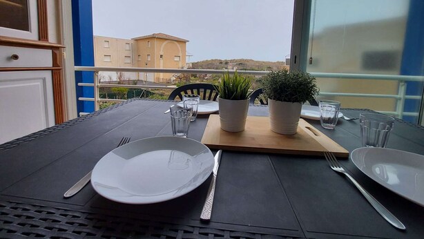 A dining table set with plates, cutlery, and glasses, with potted plants and a view of buildings in the background. - Charming 2-Room Cabin Apartment, 50m from Beach, A/C, Parking (Marseillan-Plage)