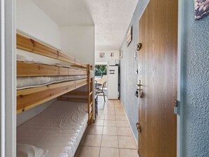 A narrow room with a bunk bed, a wooden door, and a kitchen area in the background.