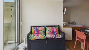 A balcony with a sliding glass door, a black sofa with colorful cushions, a small table with a chair, and a bed with a white mattress and pillows.