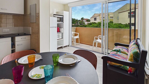 A modern kitchen with white cabinets, a dining table set with plates and glasses, and a sliding glass door leading to a balcony with chairs.