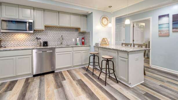 A modern kitchen with a stainless steel dishwasher, a microwave, and a herringbone-patterned backsplash. There are two bar stools and a small island with a sink. The floor has a herringbone wood pattern, and the walls are painted light blue. - Inviting Savannah Vacation Rental w/ Game Room! (Savannah)