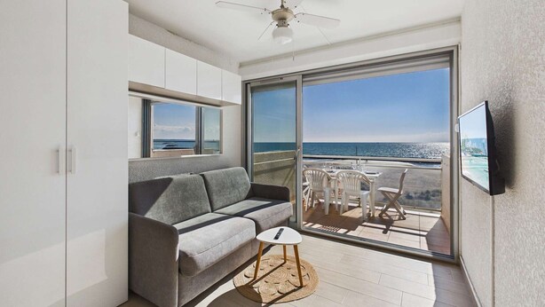 A modern living room with a gray sofa, a small round table, and a ceiling fan. Large windows offer a view of the ocean. - Grau d'Agde: Studio cabin with terrace, private parking, sea view (Le Grau d'Agde)