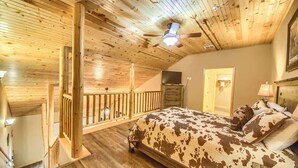 A bedroom with a wooden ceiling, a bed with a cowhide pattern, a ceiling fan, and a staircase.