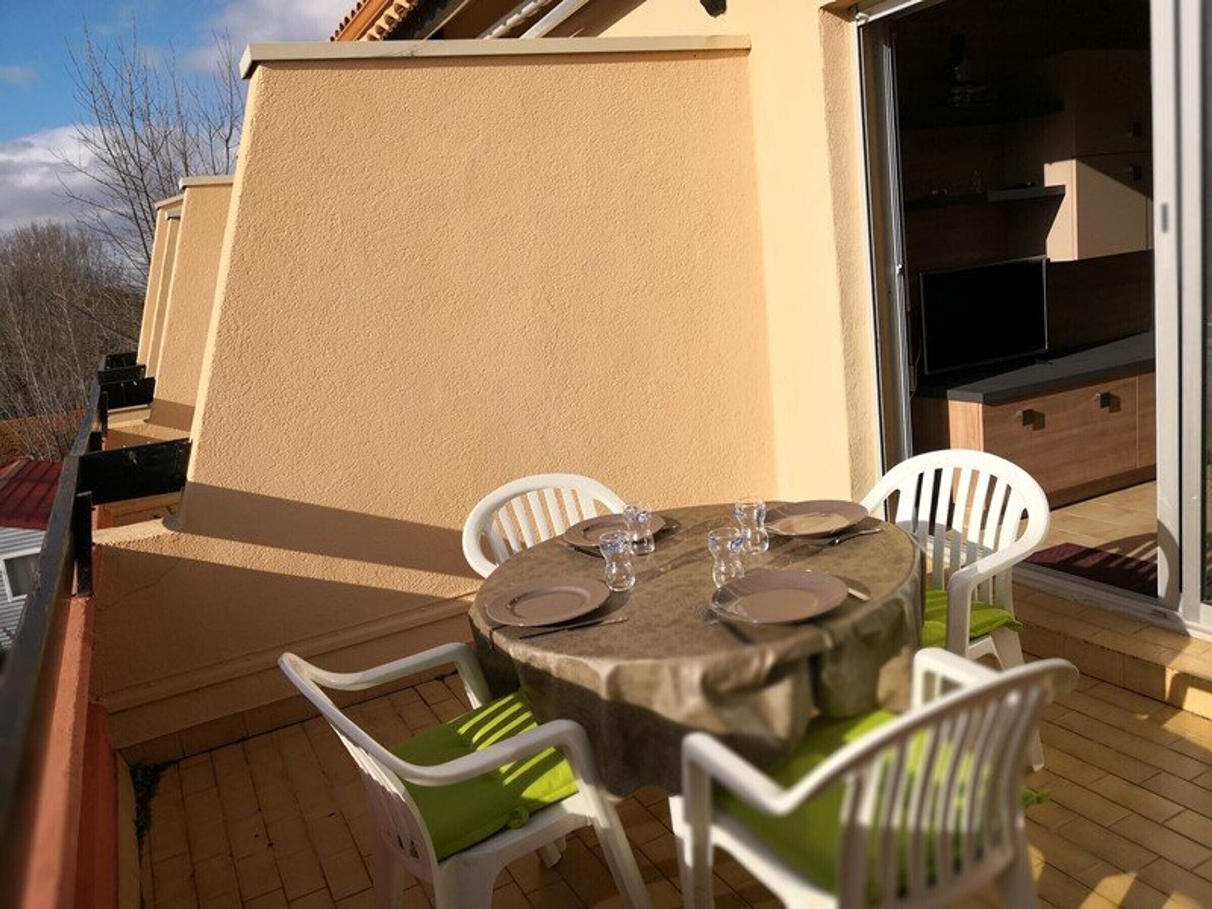Eine Terrasse im Freien mit einem runden Tisch für vier Personen, umgeben von weißen Kunststoffstühlen mit grünen Kissen.