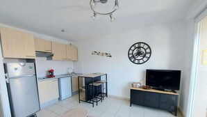 A compact living space with a kitchenette, a dining area, a sofa, a TV, and a decorative wall clock. - Beachfront 2-Room Apartment with Direct Access, Pool, and Private Parking (Marseillan-Plage)