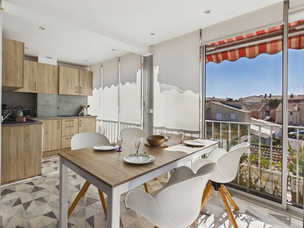 A modern kitchen with wooden cabinets, a dining area with a table and chairs, and a large window with a view of buildings.
