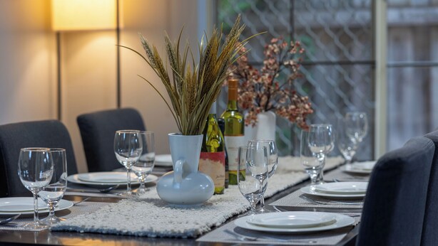 Une table à manger dressée avec des verres à vin, des assiettes et un vase décoratif contenant des plantes séchées.
