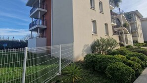 A multi-story residential building with balconies, a white fence, and well-maintained greenery.