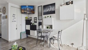 Ett kompakt bostadsutrymme med ett litet kök, en matplats med bord och stolar, en väggmonterad TV och en liten hylla med dekorativa föremål.