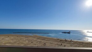 En strand med klar himmel och en skinande sol. En brygga sträcker sig ut i vattnet.