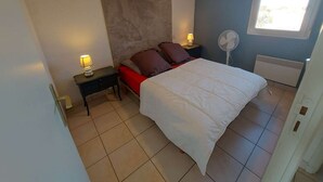 Ein Schlafzimmer mit einem Bett, zwei Nachttischen mit Lampen, einem Deckenventilator, einem Heizkörper und einem Fenster mit Tageslicht.