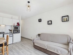 A modern kitchen with white cabinets, a dining table, and a wooden chair. There is a sofa and a small side table with a vase.