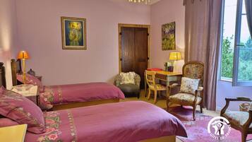 A room with two beds, a desk, and a chair. There is a window with curtains, a lamp, and a painting on the wall.
