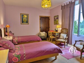 Habitación con dos camas, un escritorio y una silla. Tiene una ventana con cortinas, una lámpara y un cuadro en la pared.