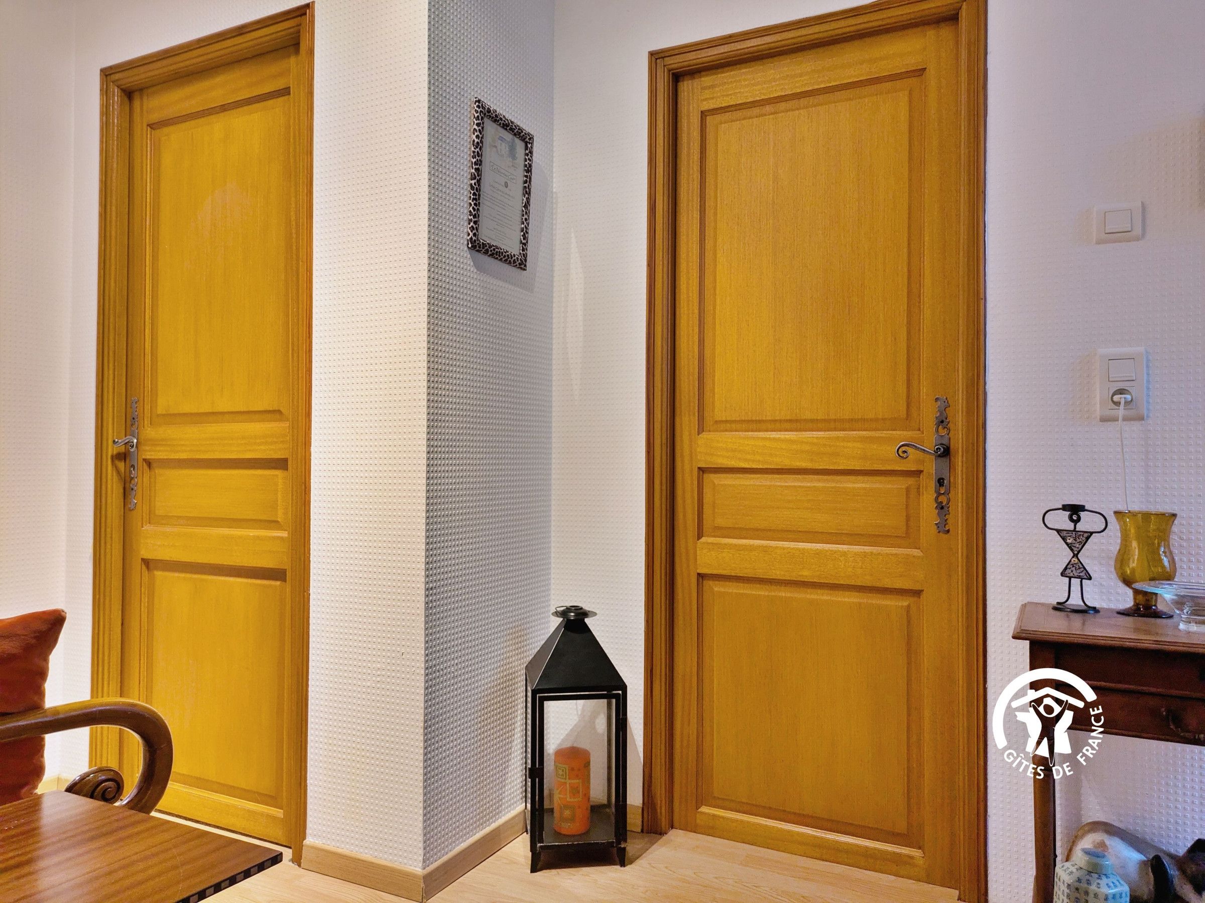 Un couloir avec deux portes en bois, un mur à texture, une petite lanterne noire et une table sur laquelle se trouve un vase.