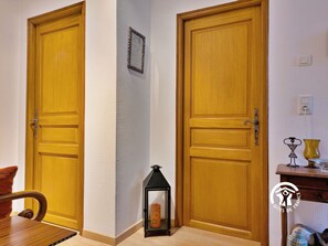 Un pasillo con dos puertas de madera, una pared con textura, un pequeño farol negro y una mesa con un florero.