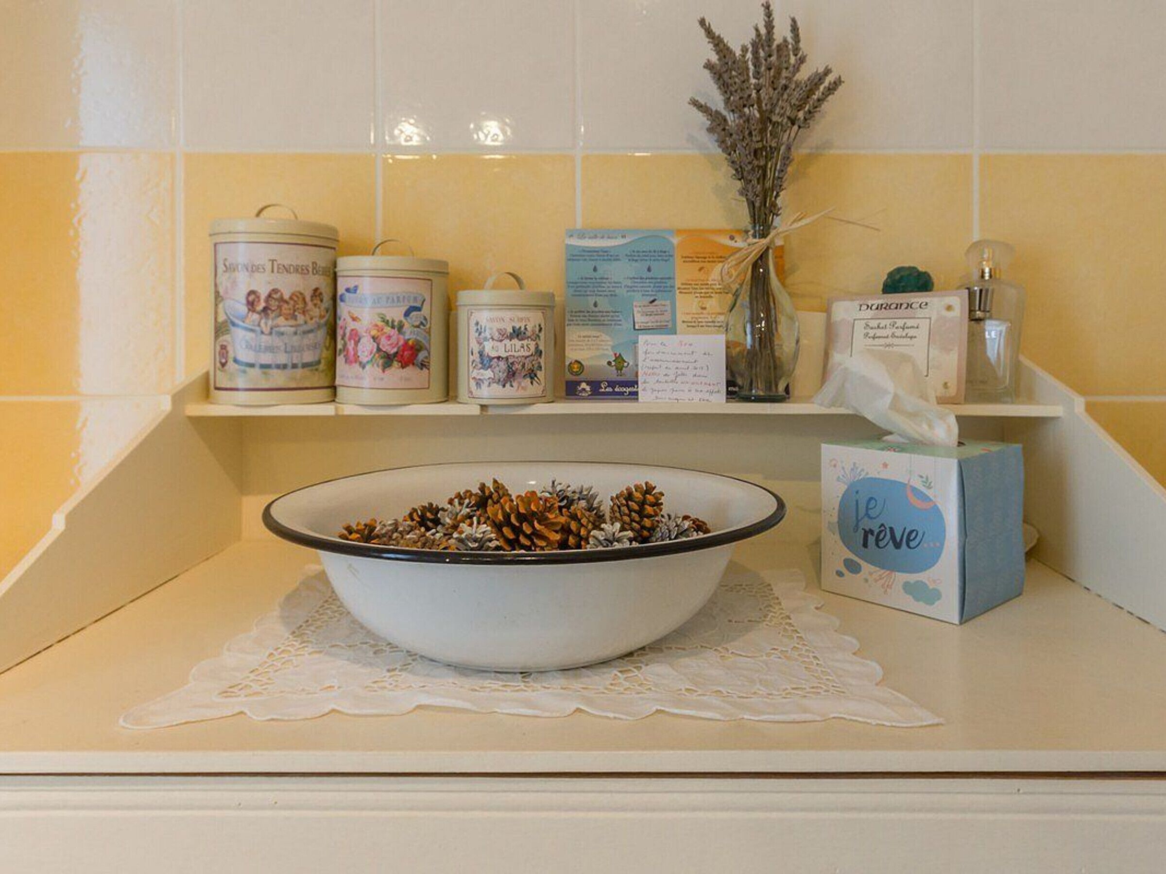 A kitchen counter with a bowl of pine cones, a box of 'je réve' soap, and several decorative items on shelves.