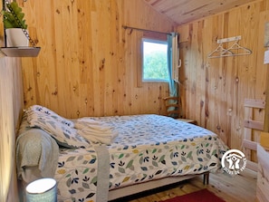 A wooden cabin room with a bed, a hanging plant, a window, and a coat rack.