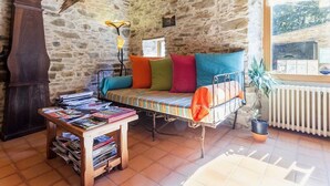 A rustic room with a stone wall, a wooden ceiling, a bed with colorful pillows, a table with magazines, a clock, and a window with a view. - Rustic Getaway: Cozy Room, Private Bath, Breakfast, and Family Friendly in Bouvron (Bouvron)