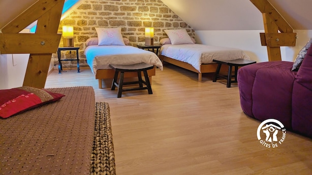 Un dormitorio cálido en el ático, con dos camas, vigas de madera, paredes de piedra y una mesita con una lámpara.
