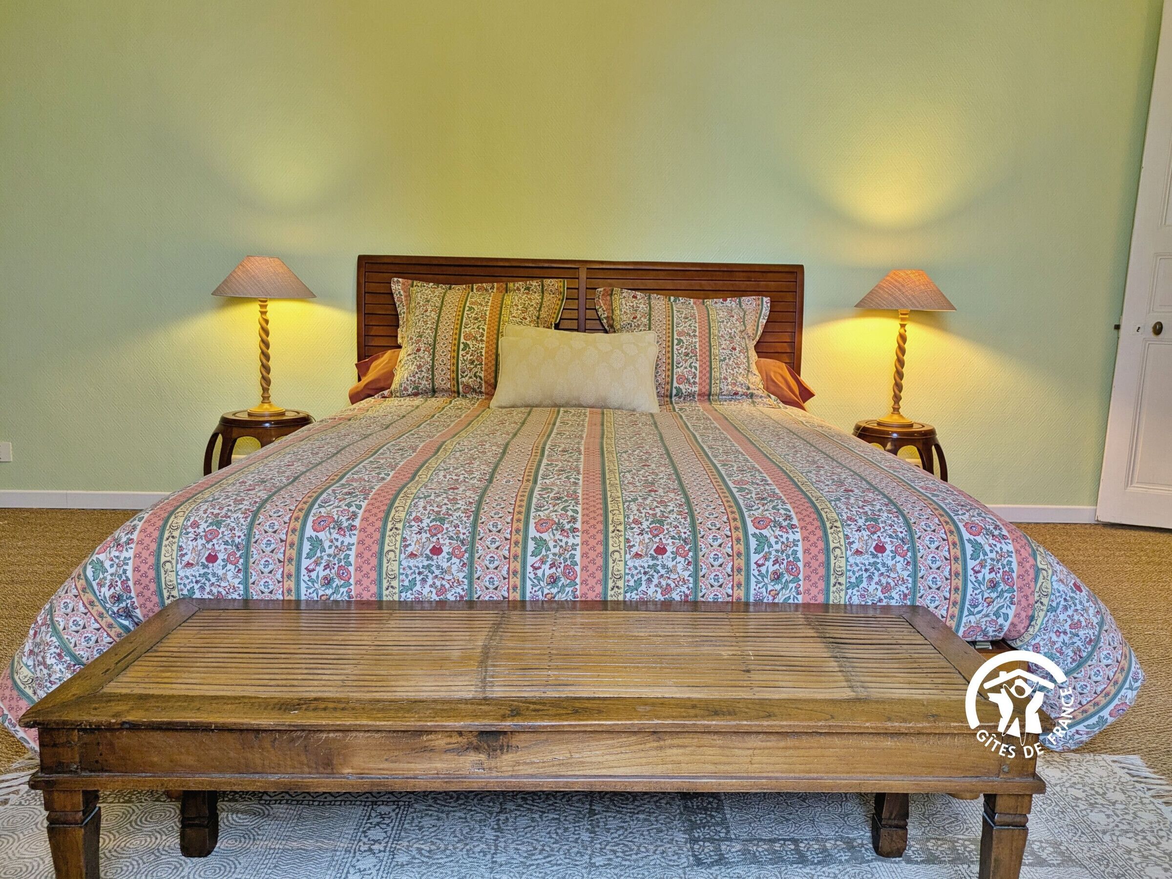 Een slaapkamer met een houten bed, bloemenbeddengoed, twee bedlampen en een houten bankje.