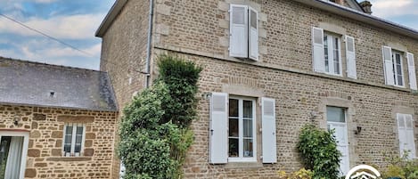 Un edificio de piedra con contraventanas blancas, un pequeño jardín y un camino de entrada de grava.